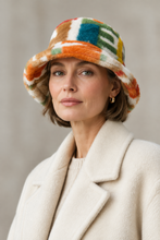 Load image into Gallery viewer, A model wearing a fuzzy bucket hat with a colorful geometric pattern in orange, green, blue, white, and brown, paired with a cream turtleneck sweater against a light gray background.
