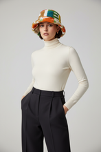 Įkelti nuotrauką į galerijos peržiūros programą, A model wearing a cream ribbed turtleneck sweater, black high-waisted trousers, and a colorful patterned bucket hat, posed against a light gray studio background.

