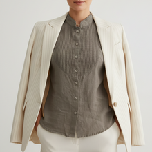 Įkelti nuotrauką į galerijos peržiūros programą, A woman stands against a light gray studio background wearing a cream pinstriped blazer over a taupe-gray pleated button-front blouse, paired with cream trousers.
