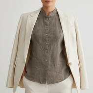 A woman stands against a light gray studio background wearing a cream pinstriped blazer over a taupe-gray pleated button-front blouse, paired with cream trousers.