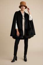 Load image into Gallery viewer, A model stands against a solid beige studio background wearing a black cape-style coat with black fur trim, a tan bucket hat, a white collared shirt, black pants, and black ankle boots.
