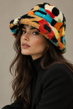 Load image into Gallery viewer, A woman wearing a fuzzy bucket hat with a vibrant multicolor geometric pattern in orange, yellow, red, blue, black, and cream, paired with a black turtleneck and blazer against a neutral beige background.
