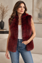 Load image into Gallery viewer, A woman modeling a sleeveless, deep burgundy faux fur vest over a white tank top and blue jeans, standing in a living room setting.
