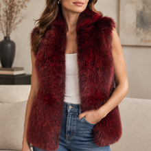 Load image into Gallery viewer, A woman wearing a sleeveless, deep burgundy faux fur vest over a white tank top and blue jeans, standing in a cozy living room setting.
