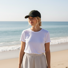 Load image into Gallery viewer, Woman standing on a beach wearing a white t-shirt and beige pants with a black cap.
