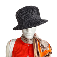 A dark grey astrakhan fur hat displayed on a mannequin wearing a red and orange outfit, with a patterned scarf.