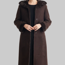 Įkelti nuotrauką į galerijos peržiūros programą, A woman wearing a long dark brown suede-like coat with a plush brown fur-trimmed hood and matching faux-fur trimmed cuffs, shown against a plain light gray studio background.
