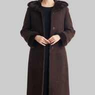 A woman wearing a long dark brown suede-like coat with a plush brown fur-trimmed hood and matching faux-fur trimmed cuffs, shown against a plain light gray studio background.