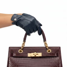 Įkelti nuotrauką į galerijos peržiūros programą, A dark navy blue leather glove on a person's hand holding the top handle of a structured brown crocodile-embossed handbag with gold hardware, against a plain white background.
