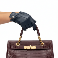 A dark navy blue leather glove on a person's hand holding the top handle of a structured brown crocodile-embossed handbag with gold hardware, against a plain white background.