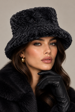 Įkelti nuotrauką į galerijos peržiūros programą, A close-up portrait of a woman wearing a black textured faux-fur bucket hat, a black fur coat, and a black leather glove, against a neutral beige background.
