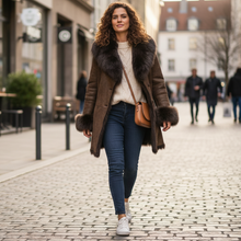 Įkelti nuotrauką į galerijos peržiūros programą, A woman walking down a cobblestone city street wearing a brown faux-fur trimmed coat over a cream sweater, blue jeans, and white sneakers, with a brown crossbody bag.
