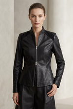 Load image into Gallery viewer, A woman modeling a black leather jacket with contrasting shoulder panels and a front zipper, paired with matching black leather pants, set against a neutral beige background.
