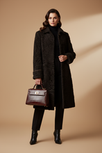 Įkelti nuotrauką į galerijos peržiūros programą, A model stands against a warm beige studio backdrop wearing a long black textured coat over a black turtleneck and black pants, holding a dark brown crocodile-embossed handbag.
