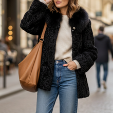 Įkelti nuotrauką į galerijos peržiūros programą, A woman stands on a cobblestone street wearing a black textured faux-fur jacket with a fur collar, a cream ribbed sweater, blue jeans, and white sneakers, while carrying a tan shoulder bag.

