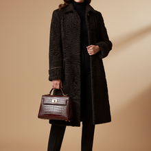 Įkelti nuotrauką į galerijos peržiūros programą, A model stands against a warm beige studio backdrop wearing a long black textured coat over a black turtleneck and black pants, holding a dark brown crocodile-embossed handbag.

