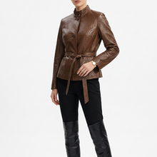 Load image into Gallery viewer, A model stands against a light gray studio background wearing a brown textured faux-leather jacket with a high collar and waist tie, black pants, and black over-the-knee boots.
