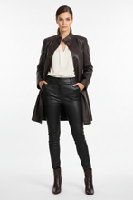 Load image into Gallery viewer, A model stands against a light gray studio background wearing a dark brown leather coat over a cream blouse, black faux-leather pants, and dark brown ankle boots.
