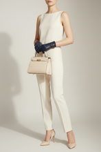 Įkelti nuotrauką į galerijos peržiūros programą, A model wearing a cream sleeveless jumpsuit, dark navy leather gloves, and beige heels, holding a structured cream handbag with gold hardware against a light gray studio background.
