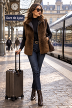 Load image into Gallery viewer, A woman stands on a Paris train station platform wearing a brown shearling vest over a black top, dark blue jeans, and brown leather boots, while pulling a brown hardshell suitcase. She is carrying a brown leather handbag.
