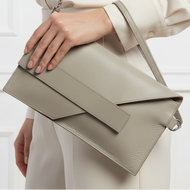 A model wearing a cream-colored satin blouse and trousers carries a structured taupe leather crossbody bag with a wide flap and thin strap.