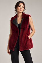 Load image into Gallery viewer, A woman modeling a sleeveless burgundy faux fur vest with black faux leather lapels, worn over a black top and black pants against a light gray studio background.
