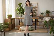 Load image into Gallery viewer, Woman in a checkered dress standing in a modern office with plants and furniture.
