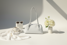Load image into Gallery viewer, White handbag on a white surface with a cup, sunglasses, and flowers.
