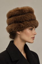 Įkelti nuotrauką į galerijos peržiūros programą, A woman in profile wearing a brown fur hat and a black coat, photographed against a plain warm gray studio background.
