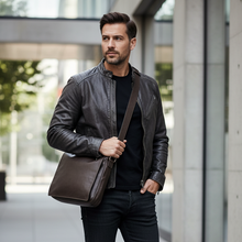 Įkelti nuotrauką į galerijos peržiūros programą, A man wearing a black leather jacket, black t-shirt, black jeans, and black boots, carrying a brown leather messenger bag with a shoulder strap while standing on a modern concrete walkway.
