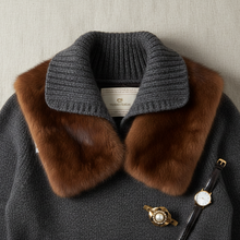 Įkelti nuotrauką į galerijos peržiūros programą, A close-up of a dark gray knit sweater with a large brown fur collar, featuring a visible VEROI GELATI brand label inside the collar. A gold-tone button with a pearl accent and a black leather watch strap are placed on the front against a neutral textured background.
