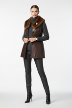 Įkelti nuotrauką į galerijos peržiūros programą, A model stands against a light gray studio background wearing a dark brown leather vest with a brown fur collar, a dark gray ribbed turtleneck, black faux-leather pants, and black heeled ankle boots.
