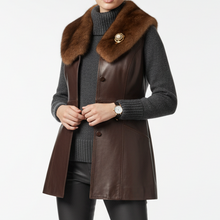 Įkelti nuotrauką į galerijos peržiūros programą, A woman wearing a dark brown leather vest with a large brown fur collar and a gold-tone decorative brooch, layered over a dark gray ribbed turtleneck sweater and black pants, against a plain white studio background.
