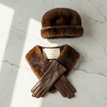 Įkelti nuotrauką į galerijos peržiūros programą, A luxury fur and leather accessory set from Moncler, featuring a brown fur hat, a matching fur collar with a Moncler label, and brown leather gloves, arranged on a white marble surface with soft window shadows.
