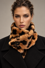 Load image into Gallery viewer, A close-up portrait of a woman wearing a plush faux-fur cowl scarf in a tan and black leopard pattern, tucked under a black coat against a neutral gray background.

