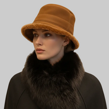 Įkelti nuotrauką į galerijos peržiūros programą, A close-up portrait of a woman wearing a warm camel-brown bucket hat with a fuzzy shearling-style trim and a large dark fur collar over a black coat, against a plain beige studio background.

