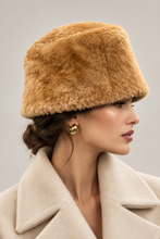 Įkelti nuotrauką į galerijos peržiūros programą, A woman in profile wearing a plush, warm camel-brown fur hat and a cream-colored coat, set against a solid light gray background.
