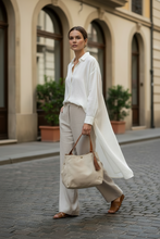 Load image into Gallery viewer, Woman in a white shirt and beige pants holding a beige handbag on a street.
