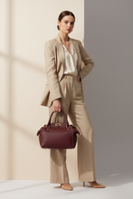 Įkelti nuotrauką į galerijos peržiūros programą, A woman models a structured burgundy leather handbag with gold hardware, styled with a beige blazer, white blouse, and beige trousers against a minimalist beige and gray background.
