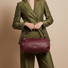 Įkelti nuotrauką į galerijos peržiūros programą, A woman wearing an olive green tailored suit holds a burgundy leather handbag with gold-tone hardware against a plain beige studio background.
