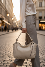 Įkelti nuotrauką į galerijos peržiūros programą, A woman in a cream blouse and light gray trousers carries a large cream-colored leather handbag with silver-tone hardware while walking down a cobblestone city street at sunset.
