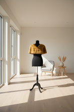 Įkelti nuotrauką į galerijos peržiūros programą, A mustard yellow faux fur cropped jacket displayed on a black dress form mannequin in a bright, modern room with large windows, a light gray armchair, and a small wooden side table.
