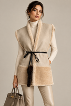Įkelti nuotrauką į galerijos peržiūros programą, A woman models a sleeveless beige and cream faux-fur vest with a black faux-leather tie belt and a dark faux-fur tassel, styled over a cream ribbed turtleneck and cream leather pants, while holding a taupe handbag against a solid light gray background.

